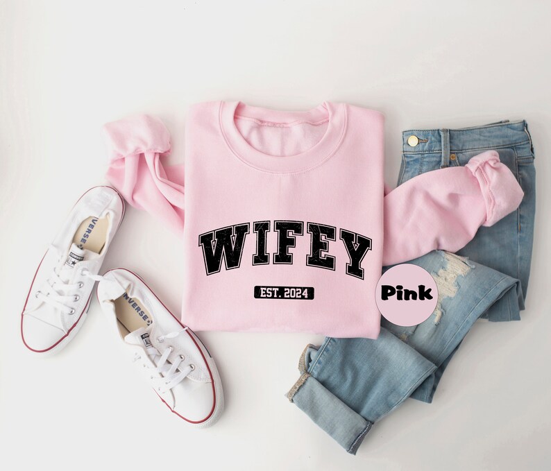 May include: Pink sweatshirt with the text "WIFEY EST. 2024" in black. The sweatshirt is laid out on a white surface with a pair of white sneakers and blue jeans.