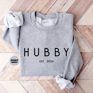 May include: A gray sweatshirt with the word "HUBBY" printed in black on the front. The text "EST. 2024" is printed below the word "HUBBY".