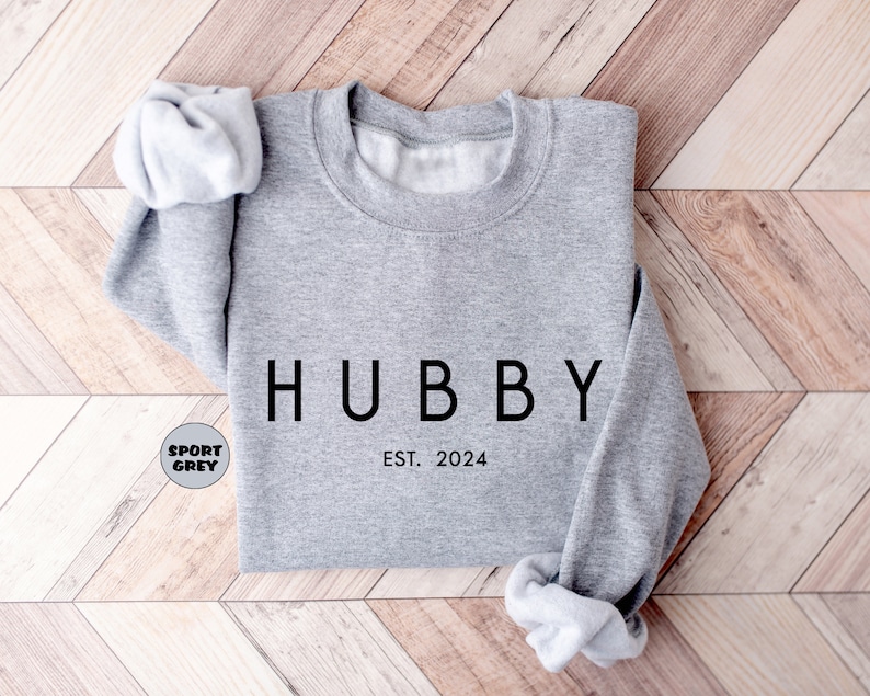 May include: A gray sweatshirt with the word "HUBBY" printed in black on the front. The text "EST. 2024" is printed below the word "HUBBY".