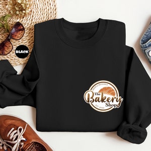 May include: Black sweatshirt with a brown and gold logo that says "The Bakery Shop" with a croissant graphic and the text "Fresh & Delicious" and "Est. 2023".
