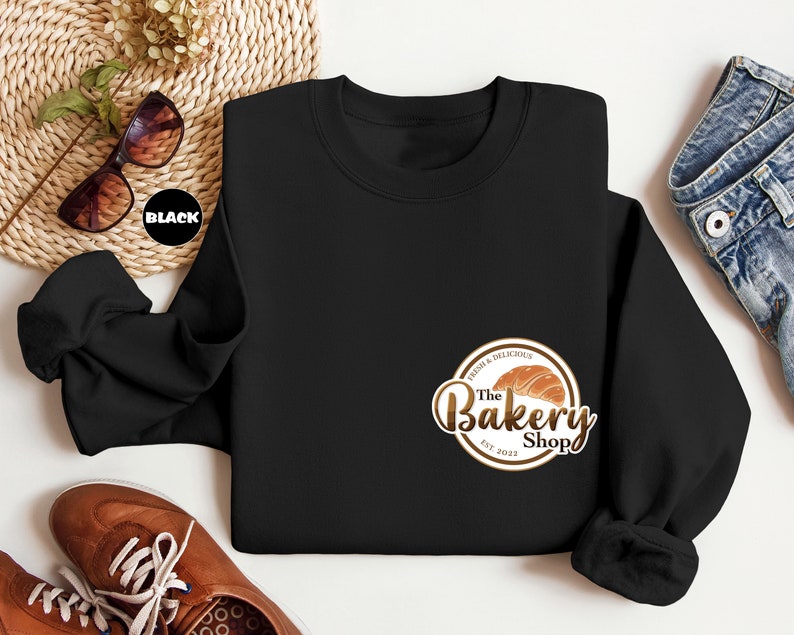 May include: Black sweatshirt with a brown and gold logo that says "The Bakery Shop" with a croissant graphic and the text "Fresh & Delicious" and "Est. 2023".