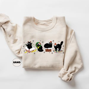 May include: A beige sweatshirt with a Christmas-themed design featuring four black cats. One cat is in a box, one is wearing a wreath, one is on a chimney, and one is holding a string of lights.