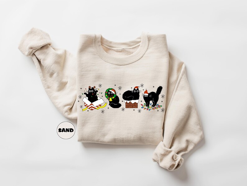 May include: A beige sweatshirt with a Christmas-themed design featuring four black cats. One cat is in a box, one is wearing a wreath, one is on a chimney, and one is holding a string of lights.