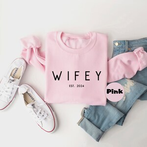 May include: Pink sweatshirt with the text "WIFEY EST. 2024" in black lettering. The sweatshirt is paired with blue jeans and white sneakers.