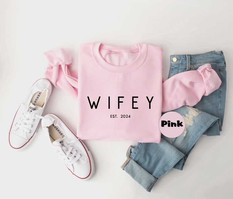 May include: Pink sweatshirt with the text "WIFEY EST. 2024" in black lettering. The sweatshirt is paired with blue jeans and white sneakers.