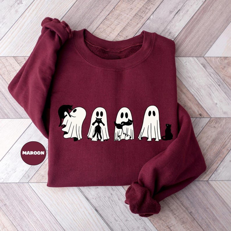 Pode incluir: Camisola de cor vinho com um gr&aacute;fico de cinco fantasmas, cada um segurando um gato preto. Os fantasmas est&atilde;o em fila, e os gatos est&atilde;o de frente para o observador. A camisola est&aacute; sobre um fundo de madeira.