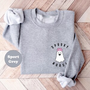 Pode incluir: Uma camisa de moletom cinza com o texto "Spooky Nurse" e um fantasma de desenho animado usando um chap&eacute;u de enfermeira rosa.