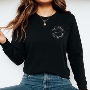 May include: Black long-sleeved top with a crew neck. The shirt has a circular logo on the upper left chest that reads "The Cuisine Restaurant" around a spoon and fork design. The shirt is worn with blue jeans.