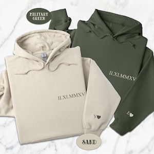 Op de afbeelding: Twee hoodies, één in zandkleur en één in militaire groen, met de Romeinse datum II.XI.MMXV en een klein hartje op elk.
