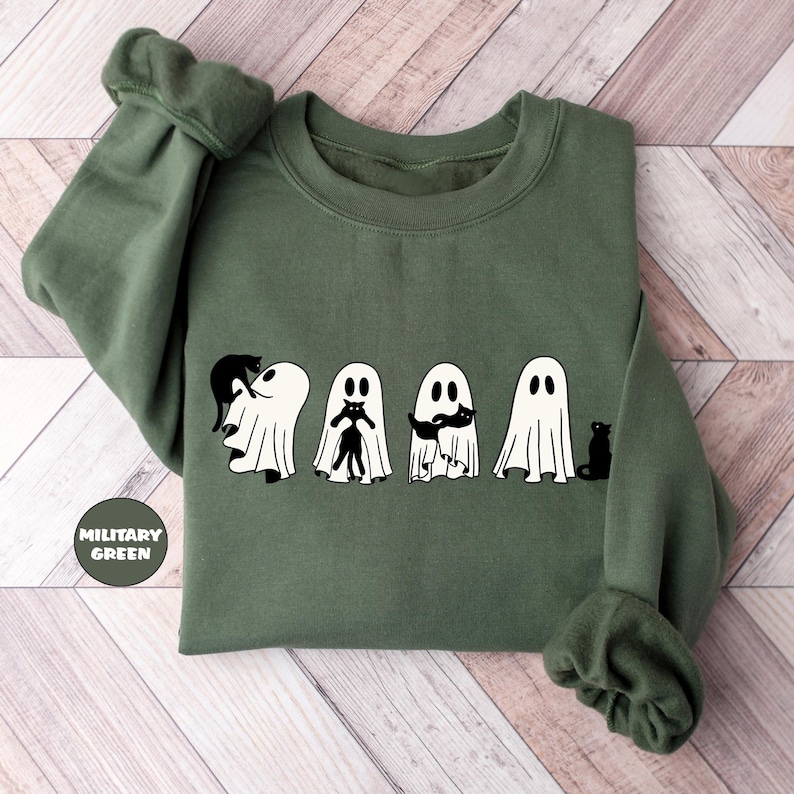 Pode incluir: Uma camisa de frio verde militar com um gr&aacute;fico de cinco fantasmas segurando gatos pretos. Os fantasmas s&atilde;o brancos e t&ecirc;m rostos sorridentes. O texto "Military Green" est&aacute; impresso no canto inferior esquerdo da camisa de frio.