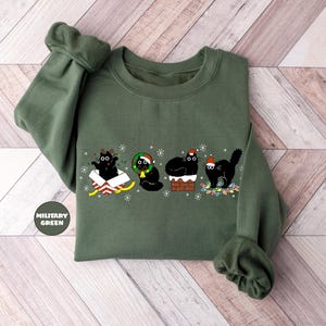 May include: A green sweatshirt with a Christmas-themed design featuring four black cats. One cat is in a box with a bow, one is wearing a wreath, one is on a chimney, and one is holding a string of lights.