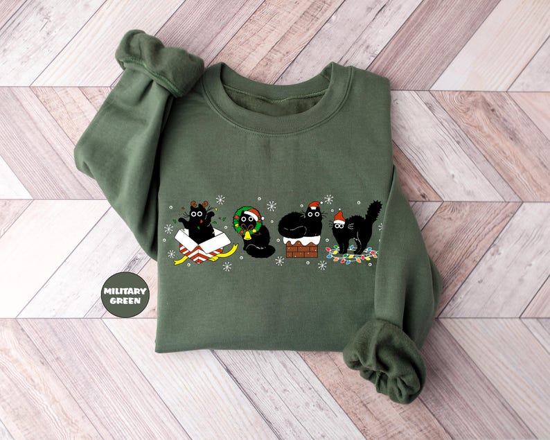 May include: A green sweatshirt with a Christmas-themed design featuring four black cats. One cat is in a box with a bow, one is wearing a wreath, one is on a chimney, and one is holding a string of lights.