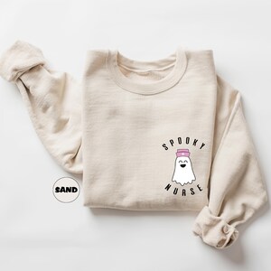 Pode incluir: Uma camisa de moletom bege com um fantasma usando um chap&eacute;u de enfermeira e o texto "Spooky Nurse" estampado na frente.