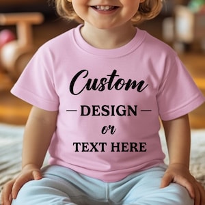 Puede incluir: Una camiseta rosa claro con las palabras "Custom - DESIGN - or TEXT HERE" en negro. La camiseta de manga corta la lleva un ni&ntilde;o.