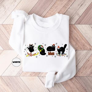 May include: White long-sleeved sweatshirt with a Christmas-themed design featuring four black cats wearing Santa hats and holding presents. The cats are surrounded by Christmas lights and snowflakes.