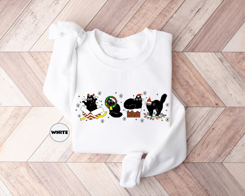 May include: White long-sleeved sweatshirt with a Christmas-themed design featuring four black cats wearing Santa hats and holding presents. The cats are surrounded by Christmas lights and snowflakes.