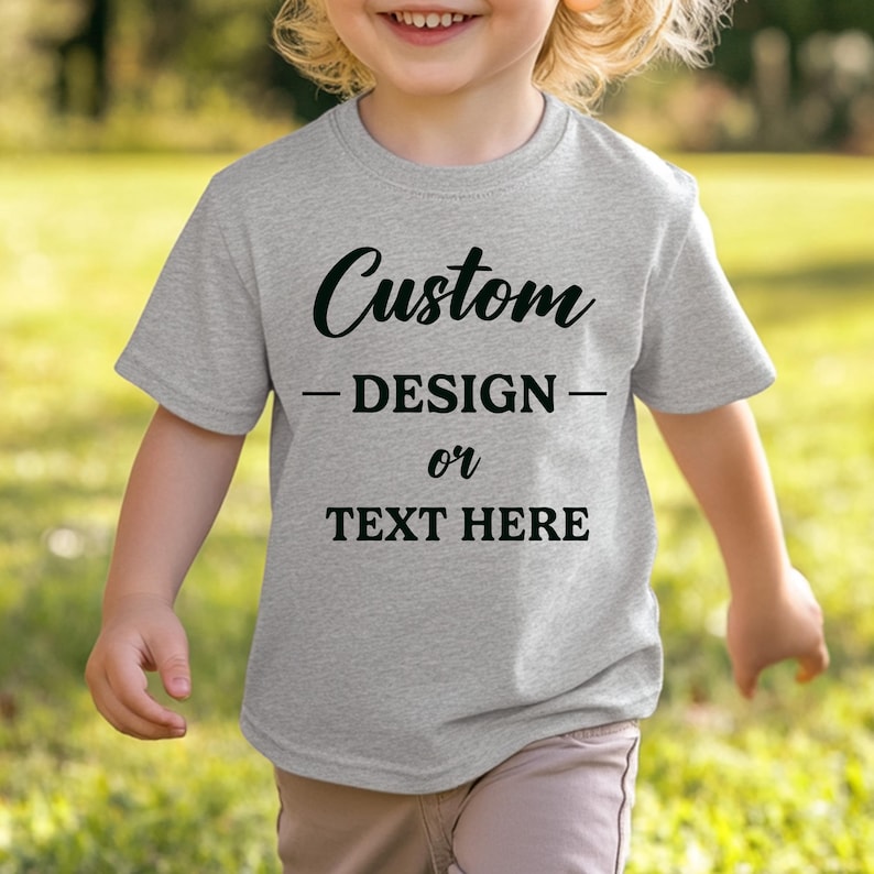 Puede incluir: Una camiseta gris claro con las palabras "Custom DESIGN or TEXT HERE" en negro. La camiseta de manga corta parece estar hecha de un material suave. La camiseta la lleva un ni&ntilde;o.
