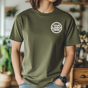 May include: Olive green t-shirt with a crew neck and short sleeves. The shirt features a white circular logo that says "YOUR LOGO HERE". The model is wearing blue jeans and a dark green watch.