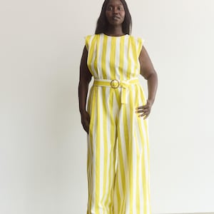 May include: A sleeveless jumpsuit featuring vertical yellow and white stripes, a round neckline, and a matching fabric belt with a circular buckle at the waist. The garment has wide-leg trousers that fall to the floor in a straight silhouette.