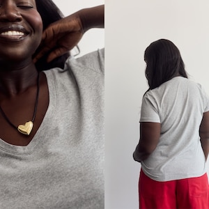 K&ouml;nnte beinhalten: Ein graues kurz&auml;rmeliges T-Shirt mit V-Ausschnitt, kombiniert mit einem goldenen herzf&ouml;rmigen Anh&auml;nger an einer schwarzen Kordelkette. Das T-Shirt ist von vorne und hinten zu sehen, wobei die R&uuml;ckansicht einen schlichten grauen Stoff zeigt.