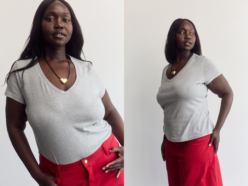 K&ouml;nnte beinhalten: Ein hellgraues kurz&auml;rmeliges T-Shirt mit V-Ausschnitt kombiniert mit einer leuchtend roten Hose. Ein goldener herzf&ouml;rmiger Anh&auml;nger h&auml;ngt an einer schwarzen Kordelkette und liegt am Ausschnitt des grauen T-Shirts an.
