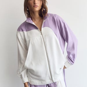 May include: A long-sleeve zip-up jacket featuring a color-blocked design in lavender and white. The jacket has a high collar, a silver-toned front zipper, and ribbed cuffs. The lavender fabric forms a curved yoke across the chest and shoulders.