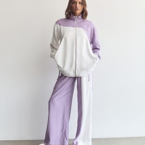 May include: A two-piece set consisting of a long-sleeve zip-up jacket and wide-leg trousers. The jacket features a color-blocked design in lavender and white with a high collar. The matching trousers are also color-blocked in lavender and white.