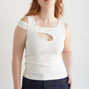 May include: A cream-coloured ribbed vest top with a distinctive cut-out detail. It has a square neckline and a single shoulder strap. Worn with dark blue denim jeans. The model is wearing gold earrings.