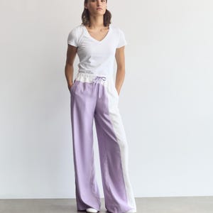 Könnte beinhalten: Ein weißes T-Shirt mit V-Ausschnitt kombiniert mit einer weiten Hose im Color-Blocking-Design. Die Hose ist vertikal in lavendelfarbene und weiße Abschnitte unterteilt, verfügt über einen elastischen Bund mit Kordelzug und hat Seitentaschen.