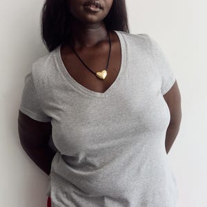 May include: A heather gray short-sleeved V-neck t-shirt paired with a gold-toned heart-shaped pendant on a thin black cord necklace and bright red pants. The t-shirt features a classic fit with short sleeves and a simple neckline design.