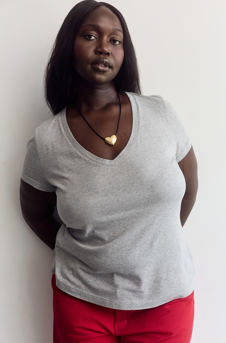 K&ouml;nnte beinhalten: Ein graues, meliertes Kurzarm-T-Shirt mit V-Ausschnitt, kombiniert mit einem goldfarbenen herzf&ouml;rmigen Anh&auml;nger an einer d&uuml;nnen schwarzen Kordelkette und einer leuchtend roten Hose. Das T-Shirt hat eine klassische Passform mit kurzen &Auml;rmeln.