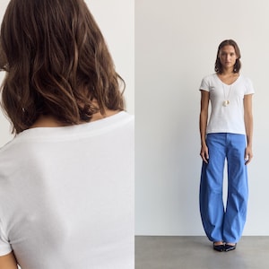 K&ouml;nnte beinhalten: Ein wei&szlig;es kurz&auml;rmeliges T-Shirt mit V-Ausschnitt kombiniert mit einer weit geschnittenen blauen Hose. Das Outfit umfasst eine lange Halskette mit Anh&auml;nger. Das T-Shirt hat ein schlichtes Design, w&auml;hrend die Hose eine lockere, weite Passform aufweist.