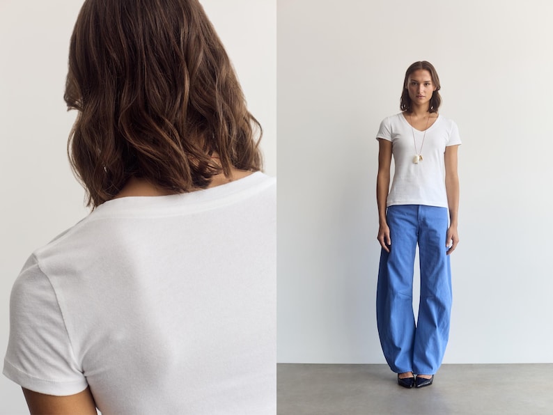 K&ouml;nnte beinhalten: Ein wei&szlig;es kurz&auml;rmeliges T-Shirt mit V-Ausschnitt kombiniert mit einer weit geschnittenen blauen Hose. Das Outfit umfasst eine lange Halskette mit Anh&auml;nger. Das T-Shirt hat ein schlichtes Design, w&auml;hrend die Hose eine lockere, weite Passform aufweist.