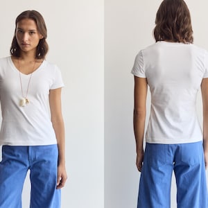 K&ouml;nnte beinhalten: Ein schlichtes wei&szlig;es kurz&auml;rmeliges T-Shirt mit V-Ausschnitt kombiniert mit einer leuchtend blauen weiten Hose. Das T-Shirt hat eine einfache, k&ouml;rpernahe Passform, w&auml;hrend die Hose einen geraden Schnitt mit sichtbaren Ges&auml;&szlig;taschen aufweist.