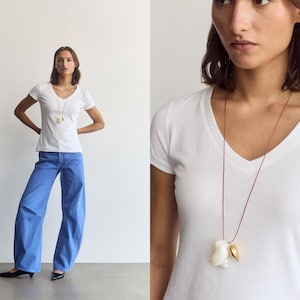 K&ouml;nnte beinhalten: Ein wei&szlig;es T-Shirt mit V-Ausschnitt kombiniert mit einer weit geschnittenen blauen Hose und einer langen Halskette aus einer d&uuml;nnen roten Kordel mit einer wei&szlig;en Muschel und einem kleinen goldenen Metallanh&auml;nger auf der Brust.