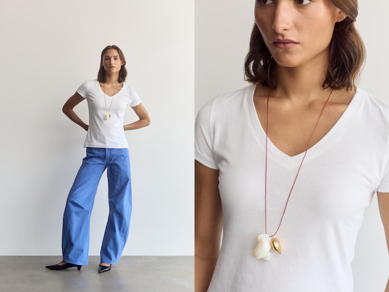 K&ouml;nnte beinhalten: Ein wei&szlig;es T-Shirt mit V-Ausschnitt kombiniert mit einer weit geschnittenen blauen Hose und einer langen Halskette aus einer d&uuml;nnen roten Kordel mit einer wei&szlig;en Muschel und einem kleinen goldenen Metallanh&auml;nger auf der Brust.