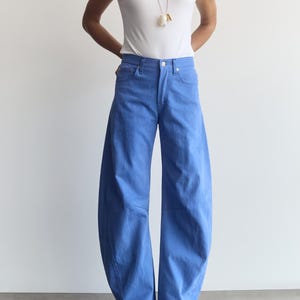 May include: A white short-sleeved V-neck t-shirt paired with bright blue wide-leg denim jeans featuring a high-waisted fit and a curved, voluminous silhouette. A long necklace with a white pendant hangs over the shirt, and black pointed-toe shoes.