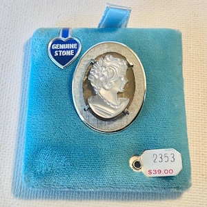 May include: An oval brooch featuring a white carved cameo profile of a person set against a dark background within a textured silver-tone metal frame. A blue heart-shaped tag reads GENUINE STONE, and a white tag displays the number 2353.