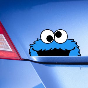 May include: A blue car with a cartoon character sticker on the back. The sticker is a blue monster with large white eyes and a black mouth. The car is parked in a parking lot.