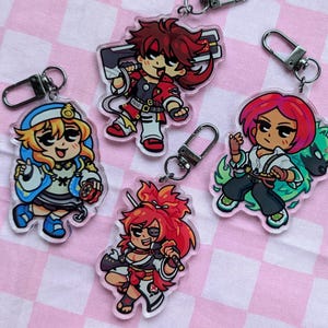 May include: A collection of acrylic keychains featuring colorful anime-style characters. Each keychain has a silver clasp for easy attachment. The characters have vibrant hair and clothing, set against a pink and white checkered background.