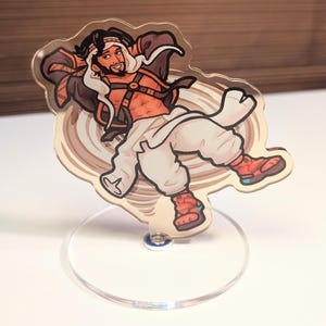 May include: An acrylic character standee featuring a cartoon illustration of a person with a beard, wearing a white head covering, a dark open jacket, and white pants, reclining against a swirling beige background, mounted on a clear circular base.