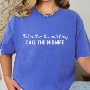 May include: A blue t-shirt with the text "I'd rather be watching CALL THE MIDWIFE" in white. The shirt is a solid color and has short sleeves. The model is wearing blue jeans.