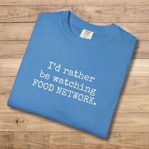 May include: A light blue t-shirt with the text "I'd rather be watching FOOD NETWORK." The shirt is folded and lies on a wooden surface. The text is in white, contrasting with the shirt's color.