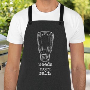 May include: A black apron with a white salt shaker illustration and the text "needs more salt." The apron is being worn by a person in front of a grill.