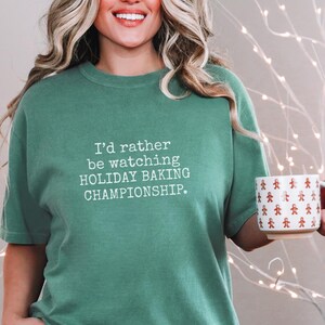 May include: A green t-shirt with the text "I'd rather be watching HOLIDAY BAKING CHAMPIONSHIP." A white mug with a gingerbread man pattern is held in the hand. The shirt is paired with blue jeans.