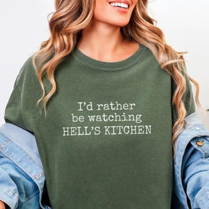 Op de afbeelding: Olijfgroene crewneck sweatshirt met de witte tekst "I'd rather be watching HELL'S KITCHEN". De sweatshirt wordt gecombineerd met een spijkerjasje en een blauwe jeans. Een plant is zichtbaar op de achtergrond.