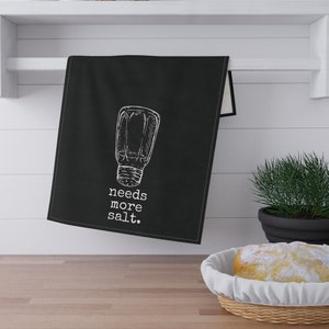 Op de afbeelding: Zwarte keukendoek met een witte illustratie van een zoutvaatje en de tekst "needs more salt."