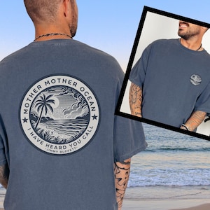 May include: Denim blue t-shirt with a circular graphic on the back featuring a palm tree, ocean, and the text "Mother Ocean, I Have Heard You Call." A smaller graphic is on the front. The shirt is a relaxed fit.