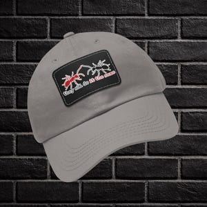 Op de afbeelding: Lichtgrijze baseballpet met een zwart embleem met twee rode en witte mierenillustraties en de tekst "they add do it the same". De pet heeft een gebogen klep.