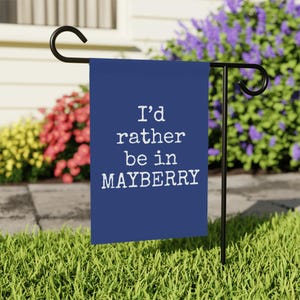 以下が含まれることがあります： 「I'd rather be in MAYBERRY」の白い文字が入った、濃いネイビーブルーのガーデンフラッグ。旗は、草が生い茂る庭の黒い金属製のスタンドに表示され、背景には色とりどりの花が咲いています。